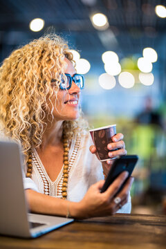 Freelance woman working remotely in a cafe, smiling while holding a coffee cup and smartphone beside her laptop, enjoying flexible work and a relaxed creative lifestyle now.