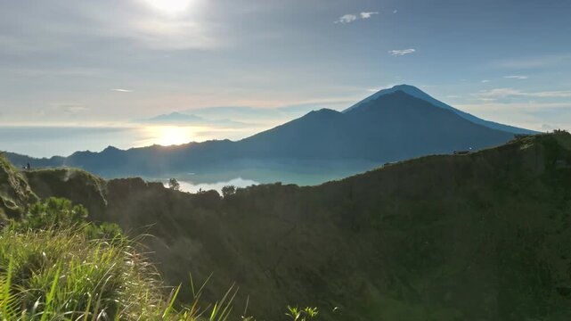 Scenic view of Mount Batur volcano and Lake Batur with rising sun illuminating the sky, creating a beautiful natural landscape with green grass in the foreground. Travel and explore Bali, Indonesia