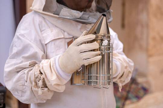 Close-up shot of a beekeeper wearing a white protective suit and thick gloves while holding a stainless steel bee smoker tool