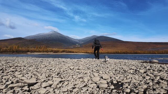 A man walks with a dog on a leash by a river. He throws stones into the water while the dog watches. The landscape has mountains and a clear sky.