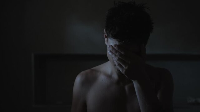 Young shirtless man covers face with hand in dim indoor interior, head bowed and shoulders hunched, conveying distress and isolation in dramatic low light portrait