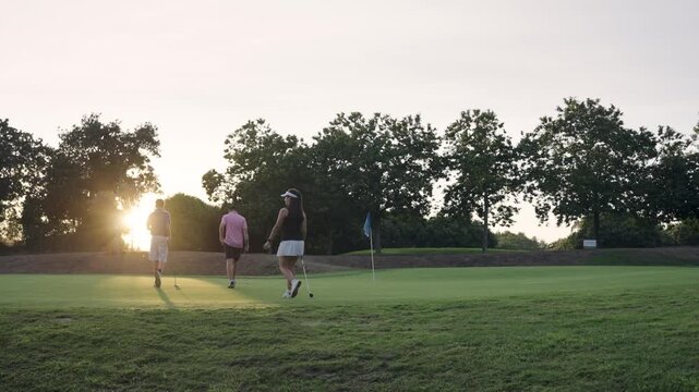Three young adult friends walking together on the golf course with putters in hand. Beautiful sunset lighting creates long shadows on the green