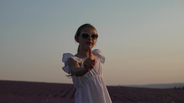 Summer fun ensues as a young girl in heart glasses dances gracefully within a blooming lavender field. Medium close-up static shot.