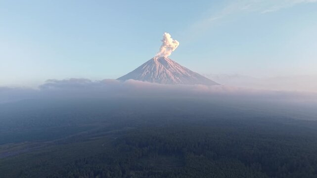 Volcano Mount Semeru erupts, smoke rises into clear sky. Dense forest blankets rolling hills beneath at Teras Semeru