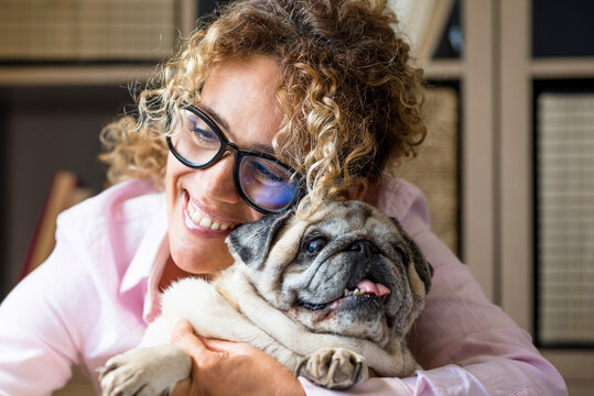 Woman with curly hair and glasses hugging a happy senior pug, smiling warmly while cuddling her pet dog, conveying affection, comfort, companionship and a joyful domestic moment.