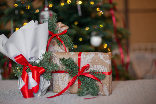 Wrapped Christmas gifts with red ribbons and pine branches in front of decorated Christmas tree. Festive spirit of Christmas giving, atmosphere of anticipation, home comfort, holiday celebration.