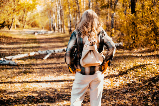 Back view of woman in autumn nature with stylish backpack. Fall outdoor hiking and mental health restoration. Leaving busy city life for peaceful solitude journey through yellow forest.