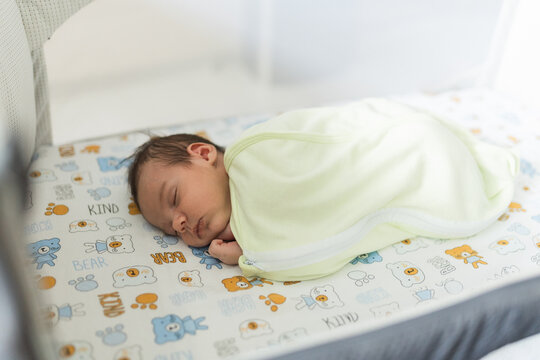 Newborn baby boy sleeping soundly inside nursery crib swaddled in soft sleeping bag.