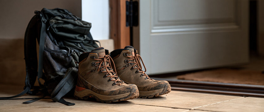 Adventure returns home mud caked hiking boots and a worn backpack rest by an open front door, evoking the satisfying end of a hard trail day. Perfect for outdoor lifestyle campaigns.