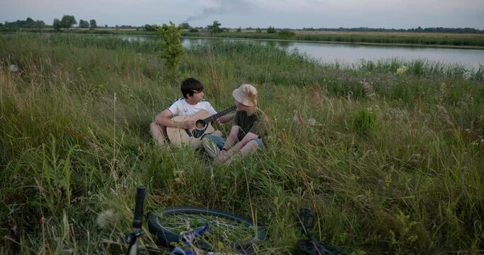 Two boys play acoustic guitar playing music enjoying nature. Teenagers spending time together. 4K 10-bit