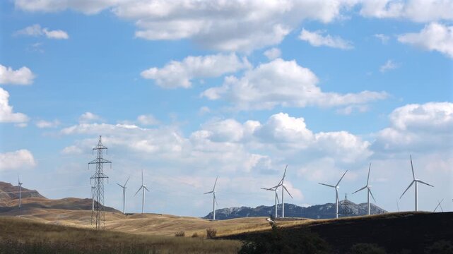 Wind Turbines and Power Lines in Mountain Landscape. Wind turbines and electricity transmission lines across a dry mountain landscape under a cloudy blue sky.