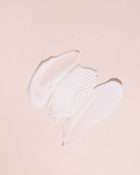 White face cream smears with wavy texture on soft peach background, moisturizer swatch close up