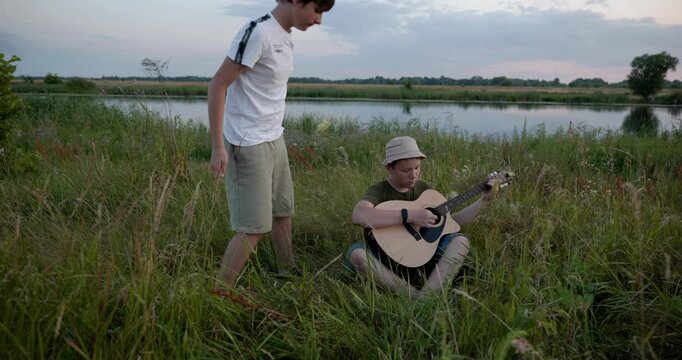 Two boys play acoustic guitar playing music enjoying nature. Teenagers spending time together. 4K 10-bit
