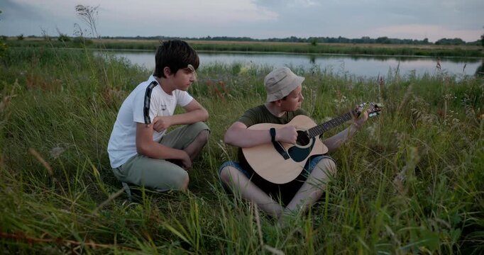 Two boys play acoustic guitar playing music enjoying nature. Teenagers spending time together. 4K 10-bit