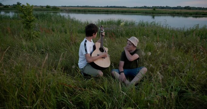Two boys play acoustic guitar playing music enjoying nature. Teenagers spending time together. 4K 10-bit