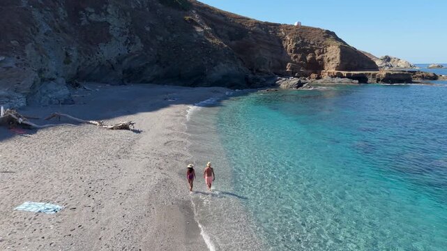 A diverse couple of man and woman walk along the sandy shoreline of La Frana Beach in Sardinia, Italy. The crystal-clear water sparkles under the sunlight, creating a perfect holiday atmosphere.