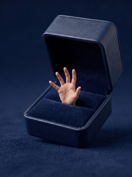 Tiny human hand reaching out from an open blue velvet jewelry ring box, surreal marriage proposal and commitment concept.