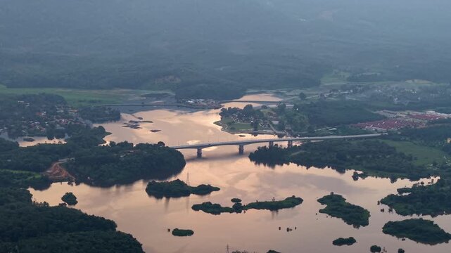 Aerial view shows a river with a bridge at golden hour. Calm waters reflect nearby hills and trees at Tasik Raban