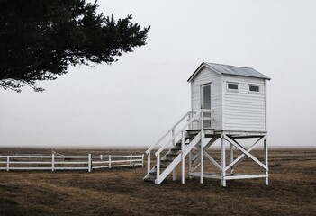 A solitary white wooden watchtower stands alone in a misty desolate field with a white fence © Png  stock