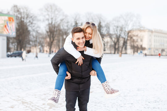 A joyful young couple enjoys a piggyback ride in a snowy city park. The woman is laughing and smiling on the man's back, wearing winter clothes. Happy date, fun mood.