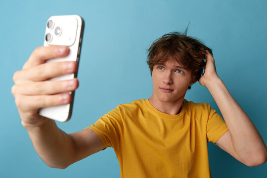 Young man in yellow t-shirt takes a selfie on smartphone, hand ruffling messy hair, hesitant gaze, searching for a good angle against blue background, relatable social media humor.