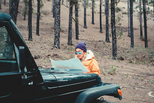 Woman in orange jacket studies a map on a black jeep hood while stopped in a pine forest, planning an outdoor road trip route during a chilly, reflective off-road adventure.