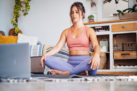 Young woman practices online yoga and guided meditation at home, sitting cross-legged on a mat while following a laptop, a calm focused fitness routine in a cozy living room.