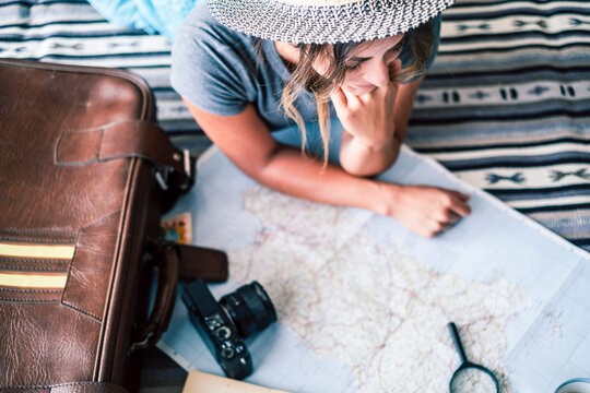 Young woman planning a trip on a paper map while lying beside a leather suitcase and camera, wearing a straw hat and casual clothes, thoughtfully tracing routes and travel ideas.