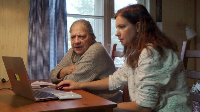 A daughter guides her elderly mother using a laptop at a wooden table in a cozy home. The concerned mother watches with arms crossed while her helpful daughter stays focused on the screen.