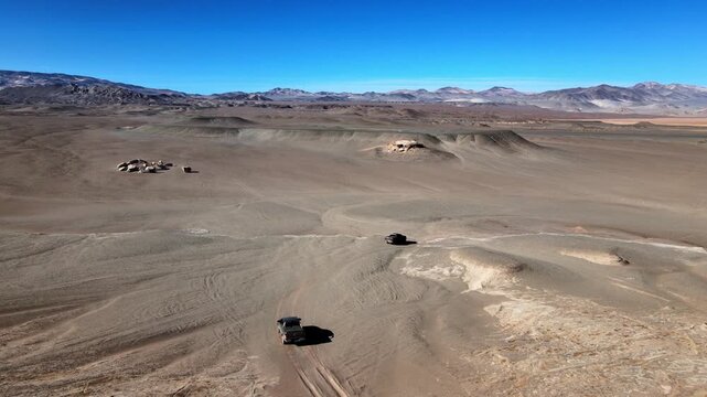 Off-road SUVs cross dry pumice plains in Campo de Piedra P&oacute;mez with distant Andean mountains under clear blue sky, useful for themes of adventure travel, remoteness and exploration.