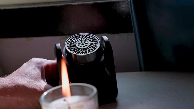 A close up of a black portable diffuser emitting white vapor behind a lit glass candle on an indoor surface. The moody scene features a bright warm flame and dark room background.