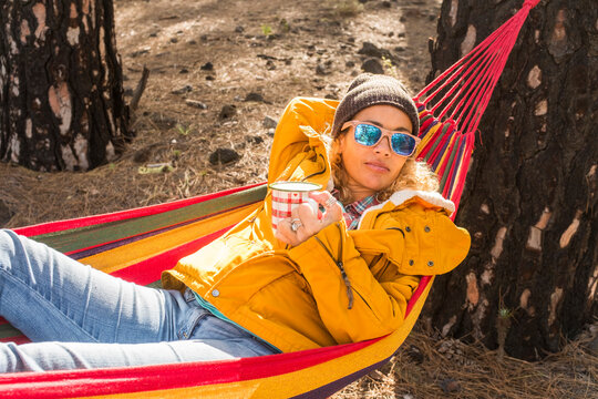 Woman in a yellow jacket lounges in a colorful hammock in a pine forest, wearing sunglasses and a beanie while sipping from a mug, enjoying a relaxed autumn outdoor camping moment.