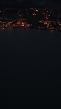 Vertical - Glowing Lights Of Lakeside Village Reflecting On Dark Waters Of Lake Achensee At Night In Tyrol, Austria. aerial tilt-up shot