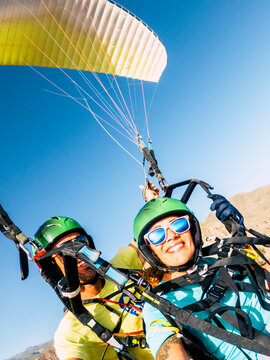 Tandem paragliding over mountains with a smiling woman in a green helmet and sunglasses, instructor behind, colorful wing overhead, sky evoking thrill, freedom and adventure.