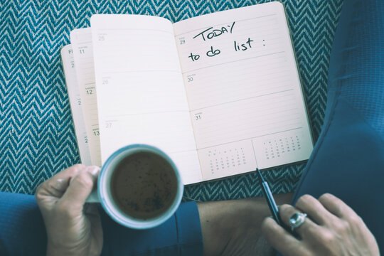 Person writing a to-do list in an open planner while holding a cup of coffee, sitting on a patterned blanket and planning daily tasks during a quiet, relaxed morning routine.