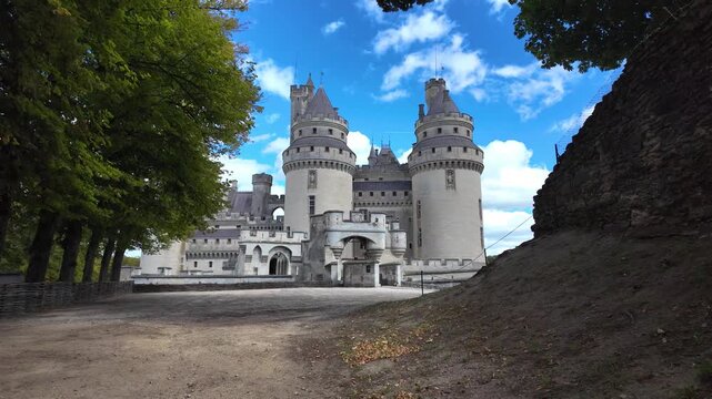Wide view of Ch&acirc;teau de Pierrefonds with twin towers, battlements and entrance structures framed by woodland, stone ramparts and a dirt pathway