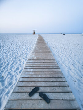 Empty wooden boardwalk across white sand to calm blue sea at dusk, with black flip-flops in foreground, conveying solitude, summer travel, minimalism and peaceful coastal mood.