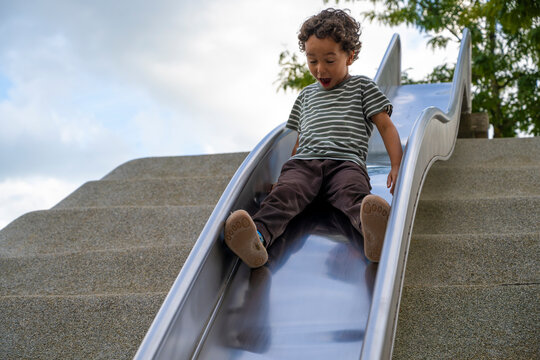 Excited young child sliding down a playground slide, enjoying active outdoor play and childhood adventure in a green park surrounded by trees and bright open sky