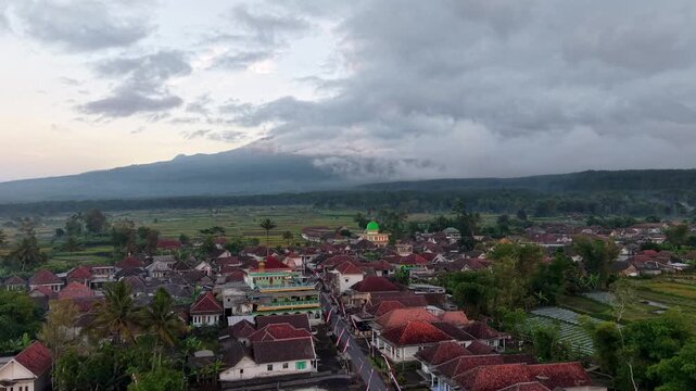 Dense clouds loom over distant mountains. Red tiled roofs dot a quiet village at Teras Semeru