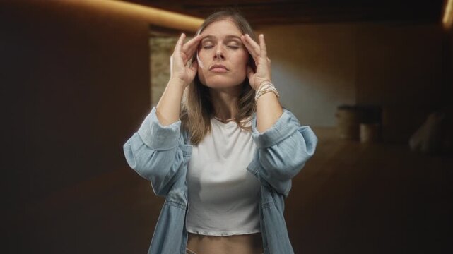 Woman presses temples inside empty studio room in blue denim shirt showing headache expression; tension discomfort.
