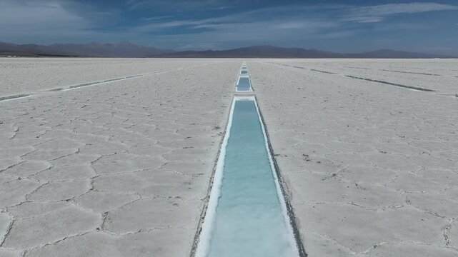 Aerial view of turquoise water channels in the vast salt flats of Salinas Grandes, Salta and Jujuy, Argentina. An endless landscape of white salt crusts under a blue sky.