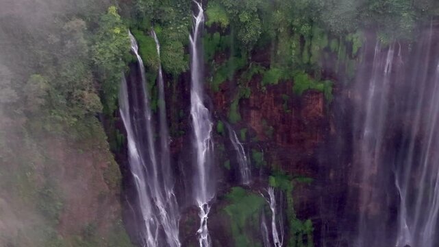 Mist veiled cascading streams fall along lush cliff face at iconic Tumpak Sewu waterfall.