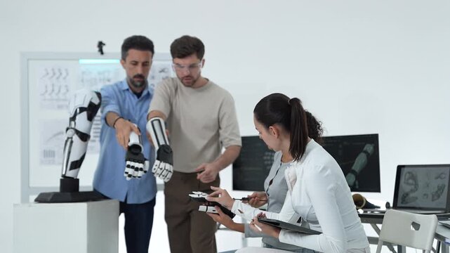 Two engineers inspecting a robotic hand beside a humanoid arm model while female researchers test a handheld bionic controller with a tablet
