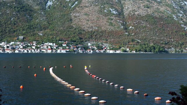 Aquaculture Farm with Floating Buoys Near Coastal Village. Rows of aquaculture buoys in calm coastal water with a small seaside village and mountains in the background.