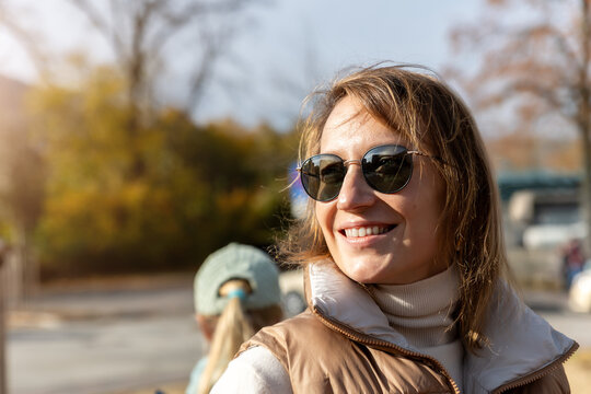 Smiling woman wearing sunglasses enjoys warm autumn sunlight outdoors in candid lifestyle portrait. Golden foliage and soft urban background create relaxed, cheerful seasonal atmosphere