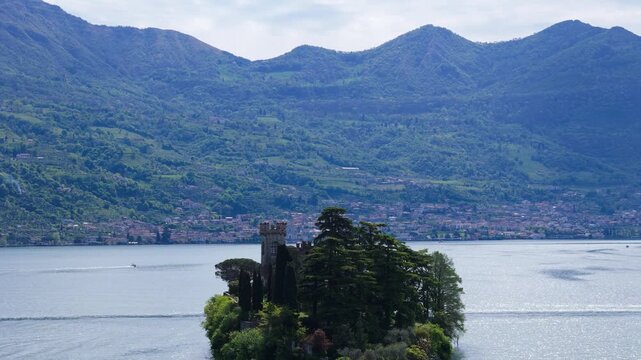 Aerial drone zoom in toward Isola di Loreto, a small private island with a historic castle surrounded by trees in the blue waters of Lake Iseo, Lombardy, Italy, with mountains in the background.