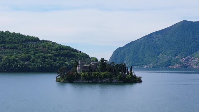 Aerial drone semi orbit around Isola di Loreto, a small island featuring a historic castle surrounded by dense trees in the blue waters of Lake Iseo, Lombardy, Italy.