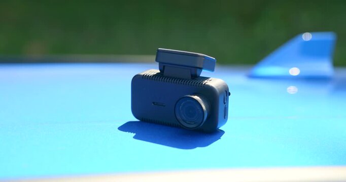 Close up of a compact black dashcam camera resting on the shiny blue roof of a car outdoors