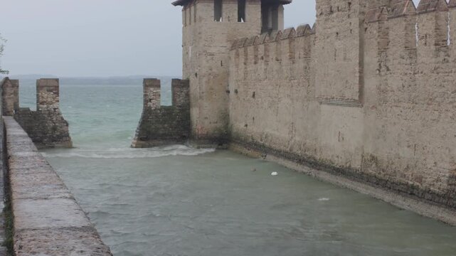 Ancient stone fortress walls at the water edge as waves break and birds glide by on a cloudy day. Tranquil medieval architecture, moody atmosphere.