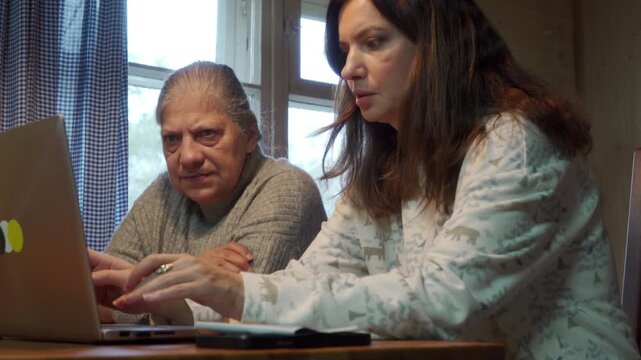 A daughter types on a laptop while her elderly mother watches attentively beside her. They sit together at a wooden table in a warm, cozy room. Their serious faces express shared concern and focus.